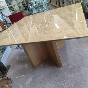 Peut inclure: Une grande table à manger carrée avec une finition en faux marbre beige clair brillant. La table présente une surface lisse et réfléchissante, soutenue par un pied central robuste composé de panneaux beiges assortis disposés en croix.