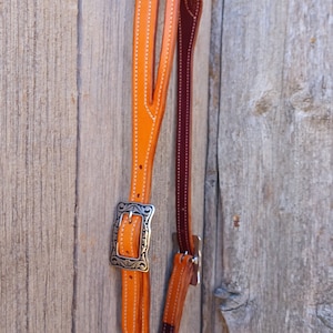 Puede incluir: Primer plano de una cabezada de cuero para caballos con una hebilla plateada. La cabezada es naranja y marrón con costuras blancas.