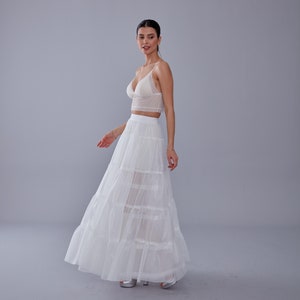 May include: A white lace crop top with a white layered tulle skirt. The model is wearing the outfit and standing on a gray background.