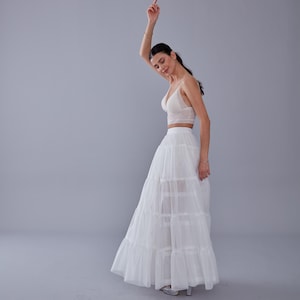 May include: A white lace crop top and a white tulle skirt with multiple layers. The skirt is long and flowing, and the model is wearing it with a casual, relaxed pose.