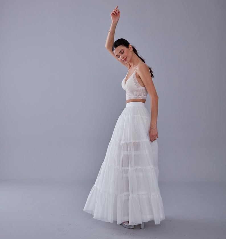 May include: A white lace crop top and a white tulle skirt with multiple layers. The skirt is long and flowing, and the model is wearing it with a casual, relaxed pose.