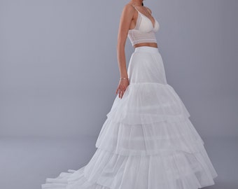May include: A white bridal ensemble featuring a lace crop top with spaghetti straps and a tiered tulle skirt. The skirt has multiple layers, creating a voluminous silhouette. The outfit is designed for a wedding or special occasion.