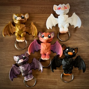 May include: Five colorful bat keychains with different colored wings and faces. The bats are made of a 3D printed material and have a shiny finish. Each bat has a silver ring attached to its back for attaching to a keychain.