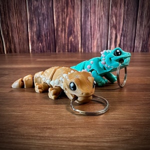 以下が含まれることがあります： 3Dプリントされた小さなゲッコーキーホルダー2個。1つは薄い茶色で黒い目、もう1つは薄い青色で黒い目。両方のキーホルダーには銀色のキーリングが付いています。