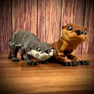 May include: Two toy otters, one gray and one brown, are posed on a wooden surface. The gray otter has a more realistic appearance, while the brown otter has a cartoonish style with large eyes and a wide smile.