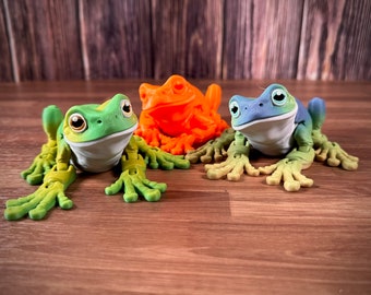 Op de afbeelding: Drie kikkerfiguurtjes, een groene, een blauwe en een oranje. De kikkers zitten met uitgestrekte poten. De figuurtjes zijn gemaakt van een glad, plastic-achtig materiaal.