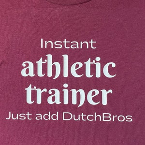 May include: Burgundy t-shirt with white text that reads "Instant athletic trainer Just add DutchBros".