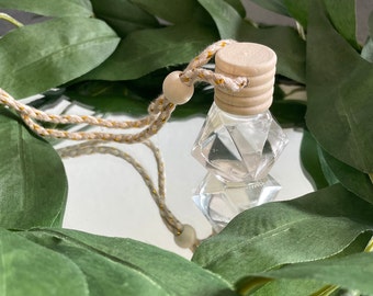 May include: A clear glass bottle with a geometric shape and a wooden lid, hanging from a beige cord with gold accents. The bottle is filled with a clear liquid and is surrounded by green leaves.