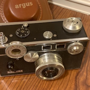 May include: A black and silver Argus camera with a silver lens and multiple dials. The camera has a black leather case with the word "argus" printed on it.