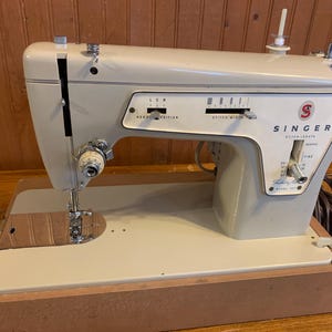 May include: A vintage Singer sewing machine in a light gray and cream color scheme. The machine features a needle position and stitch width selector, with the Singer logo prominently displayed. The machine is set in a wooden base.