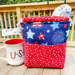 May include: A patriotic-themed yarn project bag with a red and blue star pattern. The bag has a drawstring closure and a white and gold polka dot handle. A white mug with "U.S." printed on it sits next to the bag.