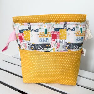 May include: A yellow fabric drawstring bag with a patchwork design featuring floral and geometric patterns. The bag has a smaller pocket on the front and a drawstring closure.