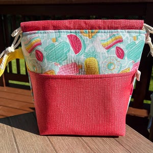May include: A fabric drawstring pouch featuring a light blue background with a colorful pattern of popsicles, donuts, and watermelon slices. The pouch has a solid red textured front pocket and a matching red top trim with white rope drawstrings.