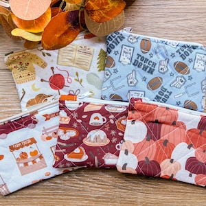 May include: Five small, quilted zipper pouches with various patterns. Designs include a sweater, open book, footballs, pumpkins, and a farm stand with pumpkins. Each pouch has a white zipper and a small tag.