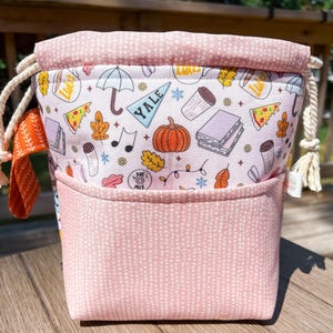May include: A pink drawstring bag with a pocket, featuring a whimsical pattern of pizza slices, pumpkins, and the word "YALE." The bag has a pink dotted pocket and a cream-colored drawstring.