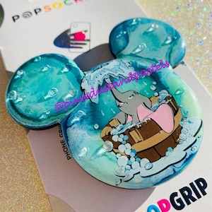 May include: A PopSocket phone grip in the shape of Mickey Mouse ears. The grip features a blue and white design with an illustration of Dumbo in a wooden tub. The PopSocket is attached to a white backing card with the PopGrip logo.