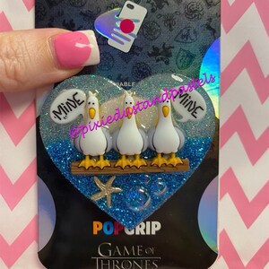 May include: A heart-shaped phone grip with a blue glitter background and four white seagulls perched on a wooden dock. The seagulls have the words "Mine" written on their backs. The grip has a gold starfish and blue bubbles. The text "PopGrip Game of Thrones" is printed below the grip.