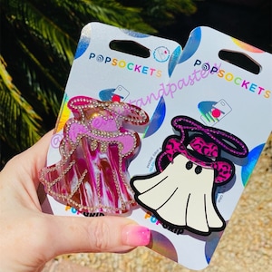 May include: Two PopSockets phone grips, one with a pink and white swirl design and a witch hat, and the other with a white ghost design with a pink and black leopard print hat.