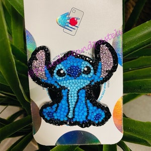 May include: A blue and black phone grip with a rhinestone design of Stitch from Lilo & Stitch. The grip is on a white card with the word "POPSOCKETS" printed on it.