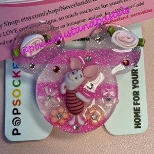 Puede incluir: Pinza para el pelo de resina rosa con purpurina en forma de orejas de Mickey Mouse con un personaje de cerdito rosa y blanco en el centro. El cerdito lleva una camisa a rayas rojas y blancas. La pinza tiene un lazo rosa con hojas verdes en la parte superior.