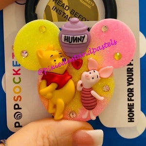 Puede incluir: Un agarre para teléfono PopSocket con temática de Winnie de Pooh y Piglet. El agarre tiene forma de orejas de Mickey Mouse. Las orejas están cubiertas de purpurina y tienen una imagen 3D de Winnie de Pooh y Piglet. El agarre tiene una olla morada con la palabra "Hunny" en ella.