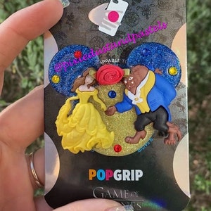Puede incluir: Un agarre para teléfono PopSocket con un diseño 3D de Bella y la Bestia de la película de Disney La Bella y la Bestia. El agarre es azul, amarillo y dorado con detalles de purpurina. El paquete incluye el texto "PopGrip" y "Game of Phones".