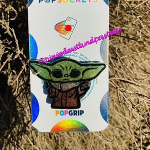 Puede incluir: Un PopGrip verde y marrón con purpurina que presenta una imagen de dibujos animados de Baby Yoda de la serie Star Wars. El PopGrip está empaquetado en una tarjeta blanca con las palabras "PopSockets" y "PopGrip" impresas en ella.