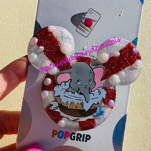 May include: A red and white glitter phone grip with a white and pink Dumbo the Elephant illustration. The grip is labeled "PopGrip" and has a white background with red polka dots. The grip is on a white card with the text "PopSockets" in rainbow letters.