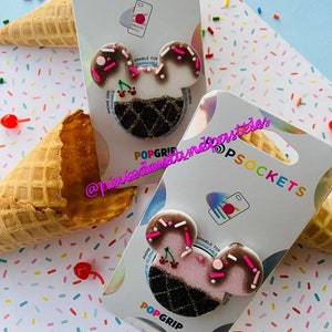 May include: Two pink and white ice cream cone shaped phone grips with chocolate and sprinkles. The grips are on white packaging with the text "POPSOCKETS" and "POPGRIP".
