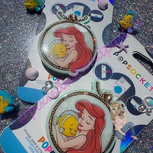 May include: Two PopSockets phone grips featuring a circular design with Ariel and Flounder from The Little Mermaid. The grips have a light blue background with a gold-colored border and a chain detail. The packaging includes the Disney logo.