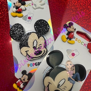 Puede incluir: Varios PopSockets con diseños de Mickey Mouse. Uno tiene pedrería negra en las orejas, otro un diseño clásico de Mickey Mouse, y otros incluyen una figura de Mickey Mouse y un guante blanco. Los PopSockets están sobre un respaldo plateado.