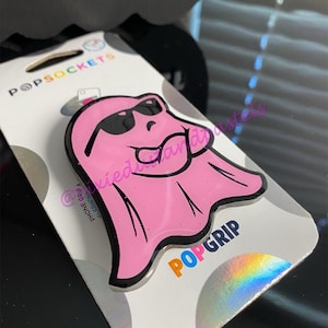 Könnte beinhalten: Ein pinkfarbener PopSocket-Telefonhalter mit einem Cartoon-Geist, der eine Sonnenbrille trägt. Der Geist hat einen schwarzen Umriss und befindet sich auf einer weißen und silbernen PopSockets-Markenrückseite. Die Worte "PopGrip" sind sichtbar.