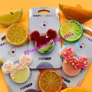 May include: A set of four phone grips featuring a Mickey Mouse design. The grips are made of clear plastic with a variety of fruit-themed designs, including watermelon, lime, orange, and a pink donut with sprinkles. The grips are all labeled "PopGrip" and are designed to be interchangeable.