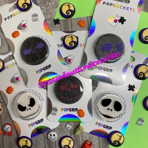 May include: Assortment of Nightmare Before Christmas themed phone grips. The grips feature different characters from the movie, including Jack Skellington, Sally, and Oogie Boogie. The grips are all black and white with a pop of color. The text "POPGRIP" is printed on each grip.
