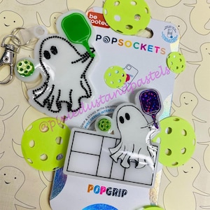 May include: A white ghost holding a green paddle with the text "be rooted" on it. The ghost is on a white background with the text "POPSOCKETS" and "GRIP & STAND WITH SWAPPA - AVEC DESSUS INTERCHA" in black. There is a white ghost holding a purple paddle with glitter on it. The ghost is on a white background with the text "POPGRIP" in rainbow colours. There are green and white pickleball paddles in the background.