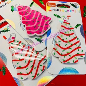 May include: Three Christmas tree-shaped phone grips with a white background and red and green designs. The phone grips are packaged on white cards with the brand name "PopSockets" printed on them.