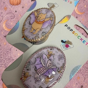 May include: Two oval PopSockets phone grips on retail packaging. One features Winnie the Pooh dressed as a bat, the other features Piglet dressed as a bat. Both are surrounded by a border of small, sparkling rhinestones and a background pattern of bats.