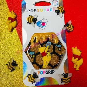 Puede incluir: Un agarre para teléfono dorado y negro con un diseño de Winnie the Pooh. Pooh está vertiendo miel de una olla en un panal. El agarre está rodeado de abejas y tiene el texto "PopGrip" en la parte inferior.