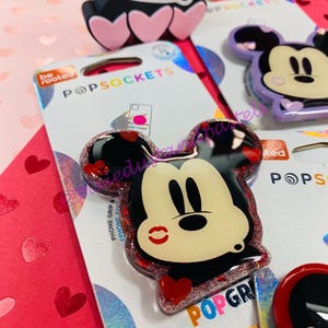 Puede incluir: Un PopSocket para teléfono de color rojo y negro con purpurina que presenta una imagen de dibujos animados de Mickey Mouse con un corazón rojo y una marca de beso roja en la mejilla. El texto "PopSockets" y "PopGrip" es visible en el embalaje.