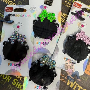 May include: Black PopSockets phone grips in the shape of cauldrons, each adorned with colorful, sparkling embellishments. One features a green design, another pink, and others with iridescent accents. The packaging includes a witch hat and the text "be rooted".