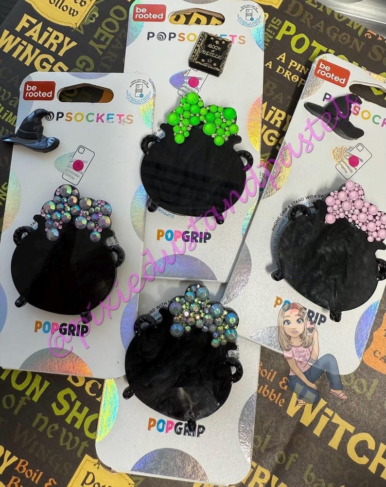 May include: Black PopSockets phone grips in the shape of cauldrons, each adorned with colorful, sparkling embellishments. One features a green design, another pink, and others with iridescent accents. The packaging includes a witch hat and the text "be rooted".