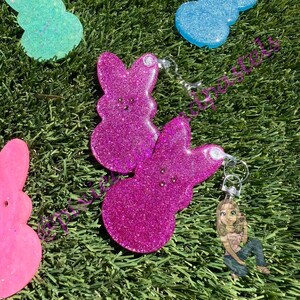 May include: A set of four glittery resin bunny keychains. The keychains are shaped like Easter Peeps and come in pink, blue, and light blue colors.