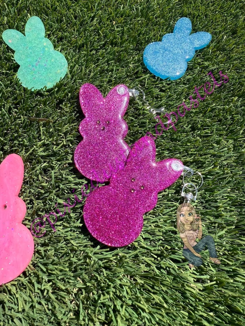 May include: A set of four glittery resin bunny keychains. The keychains are shaped like Easter Peeps and come in pink, blue, and light blue colors.