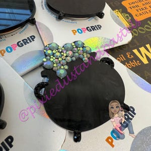 May include: Black PopGrip phone grips in the shape of a cauldron, adorned with iridescent rhinestones and a pink or green bubbling effect. The PopGrip base is black with a circular design. The packaging says "POPGRIP".