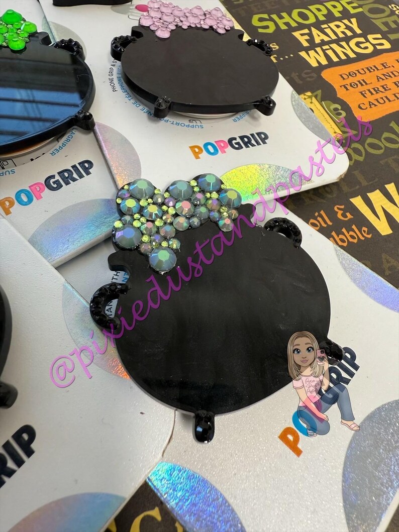 May include: Black PopGrip phone grips in the shape of a cauldron, adorned with iridescent rhinestones and a pink or green bubbling effect. The PopGrip base is black with a circular design. The packaging says "POPGRIP".