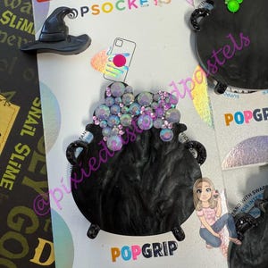 May include: Black PopSockets phone grips with a witch's hat and a cauldron design. The cauldron is embellished with iridescent rhinestones. The packaging includes the text "be rooted" and "POPGRIP".