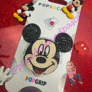May include: A PopSocket phone grip featuring a Mickey Mouse design with black and white rhinestones. The PopSocket has a swappable top and is surrounded by Mickey Mouse figurines and a cartoon illustration on a red glitter surface.
