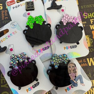 May include: Several PopSockets phone grips are displayed, featuring a black cauldron design with colorful embellishments. Other designs include a witch hat and a book. The PopSockets are on white backing cards.