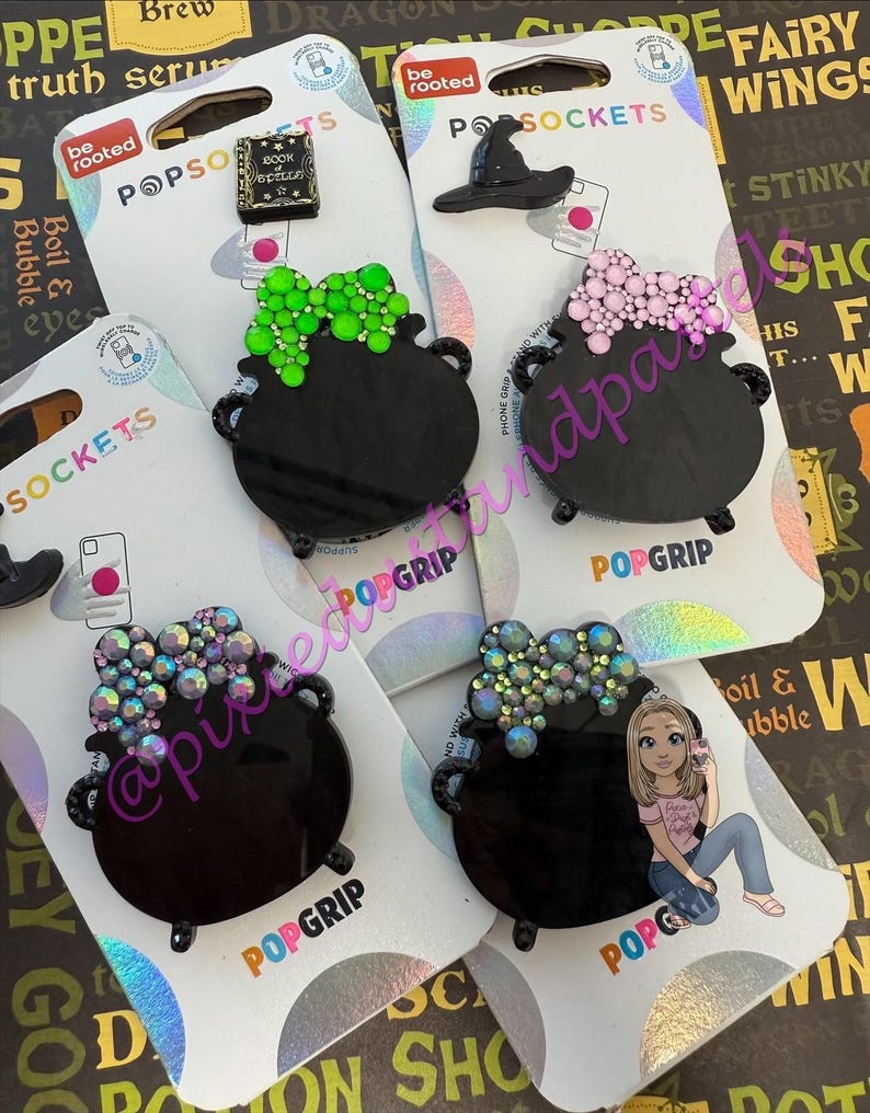 May include: Several PopSockets phone grips are displayed, featuring a black cauldron design with colorful embellishments. Other designs include a witch hat and a book. The PopSockets are on white backing cards.