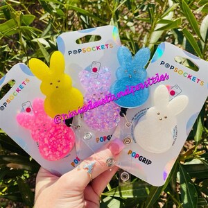 May include: Four colorful, glittery, bunny-shaped phone grips with a white background. The phone grips are attached to clear plastic badge holders with the text "POPGRIP" printed on them. The phone grips are in the colors pink, yellow, blue, and white.